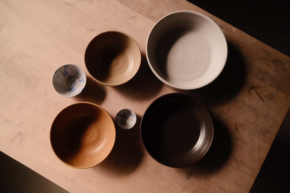 Overhead view of stoneware bowls in white, tan, blue and dark glazes arranged on a wooden table.