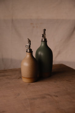 Small tan Wander ceramic oil bottle in front of a taller dark green bottle, both with metal pour spouts on a brown tabletop.