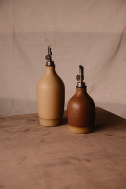 Tall tan ceramic oil bottle beside a smaller reddish-brown bottle, both with metal pour spouts on a brown tabletop.